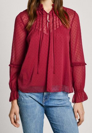 Kobieta ubrana w przezroczystą, bordową bluzkę z długim rękawem, o kropkowanej fakturze, z falbankami na mankietach i wiązaniem przy szyi, w połączeniu z niebieskimi dżinsami.