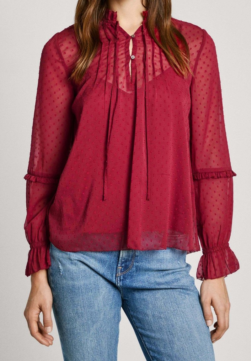 Femme portant un chemisier bordeaux transparent à manches longues avec une texture à pois, des poignets volantés et un col noué, associé à un jean bleu.