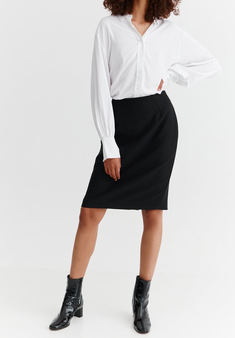 Weißes langärmliges Hemd mit Knopfleiste, kombiniert mit einem figurbetonten schwarzen Bleistiftrock und glänzenden schwarzen Stiefeletten. Glattes Material und maßgeschneiderte Passform sind sichtbar.