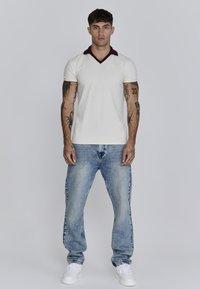 Witte polo met korte mouwen en een donker bordeaux V-hals kraag, gecombineerd met lichtblauwe distressed jeans en witte sneakers. Model staat rechtop.