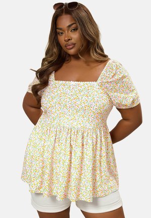 Vrouw met lang golvend bruin haar, zonnebril op haar hoofd en een wit bloesje met bloemenprint en pofmouwen, gecombineerd met een witte korte broek.