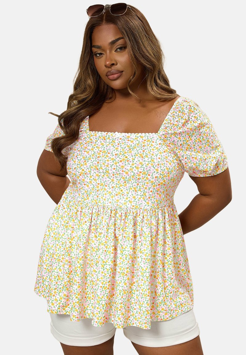 Mujer con cabello largo y ondulado de color castaño, con gafas de sol sobre la cabeza y un top blanco con mangas abullonadas y estampado floral, combinado con pantalones cortos blancos.