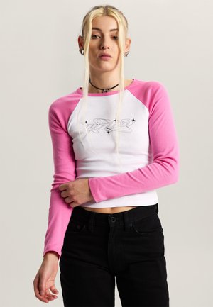 Jong persoon met lang blond haar draagt een roze-wit shirt met lange mouwen en zwarte afbeelding, zwarte broek en een choker, staand tegen een effen achtergrond.