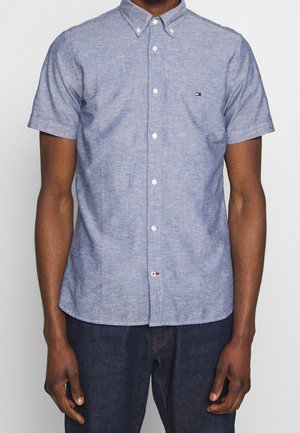 Homme portant une chemise bleu clair à manches courtes avec un petit logo sur la poitrine et un jean bleu foncé.