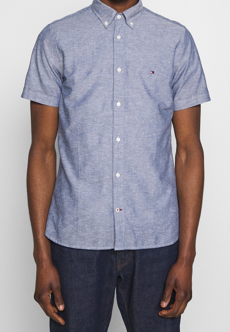 Homme portant une chemise bleu clair à manches courtes avec un petit logo sur la poitrine et un jean bleu foncé.