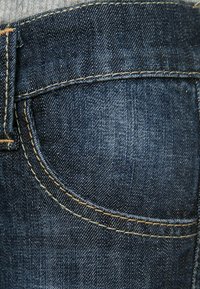 Tela de mezclilla azul oscuro que muestra un primer plano de un bolsillo trasero con costuras amarillas y una ligera textura, enfatizando la durabilidad del material.