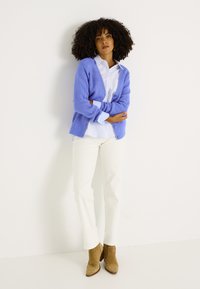 Cárdigan de punto azul claro sobre una camisa blanca a rayas, combinado con pantalones crema y botines marrones. Textura plana y tela arrugada.