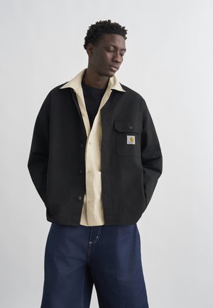 Joven vestido con camisas superpuestas de botones en negro y beige con un parche de logo, y pantalones holgados azul oscuro, posando frente a un fondo liso.