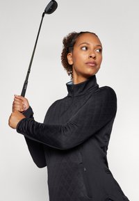 Žena držící vztyčenou golfovou holi, oblečená v černém prošívaném dlouhém rukávu, soustředěně hledí doprava na jednoduchém pozadí.