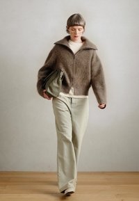 Sweater castanho com fecho éclair e gola alta, usado sobre uma blusa creme. Calças largas verdes claras e uma mala de ombro verde escuro. Textura suave.