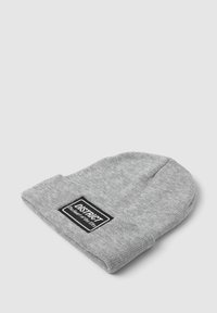 Berretto a maglia grigio chiaro con una toppa rettangolare nera che riporta la scritta "DISTRICT Il cuore della città" su uno sfondo bianco semplice.