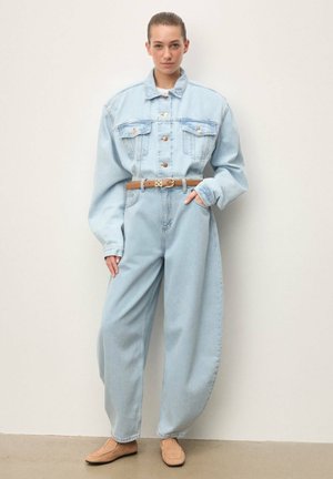 Mujer con chaqueta vaquera azul claro de gran tamaño y vaqueros anchos a juego, con cinturón marrón y mocasines beige, de pie contra una pared blanca.