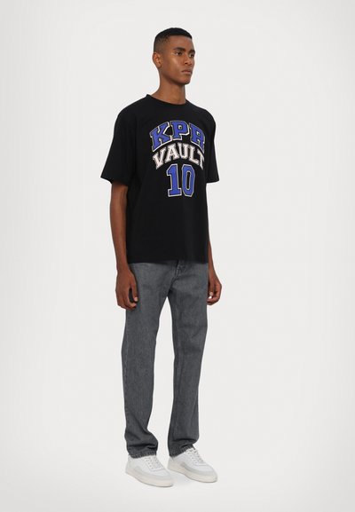Schwarzes, übergroßes T-Shirt mit "KPR VAULT 10" in blauer und weißer Schrift. Getragen mit grauen Jeans und weißen Sneakern; lässiger Streetwear-Stil.
