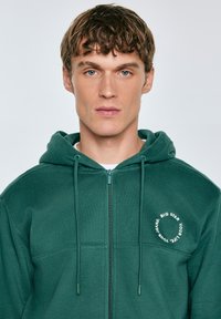 Zelená mikina s kapucí na zip, nastavitelnou šnůrkou a texturovaným povrchem. Na prsou má kruhové logo.