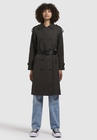 Trench coat maro închis cu nasturi dubli, talie cu curea și epauleți, asortat cu blugi albaștri deschiși și adidași negri.
