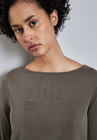 Maglietta a maniche lunghe di colore verde oliva realizzata in tessuto morbido. Presenta un collo a barchetta e testo in rilievo: "TIMELESS" con ulteriore scrittura sottostante.