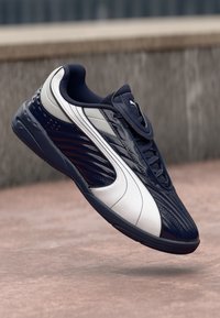 Mørkeblå og hvid Puma sneakers hængende over en belagt overflade med en struktureret stenmur i baggrunden.