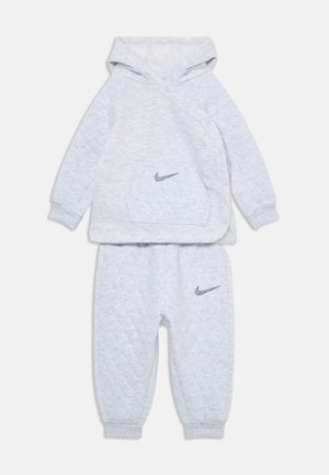 Pilkas džemperis ir kelnių komplektas, pagamintas iš struktūrinės medžiagos, su gobtuvu, priekiu kišene ir Nike logotipais ant krūtinės ir šlaunies.