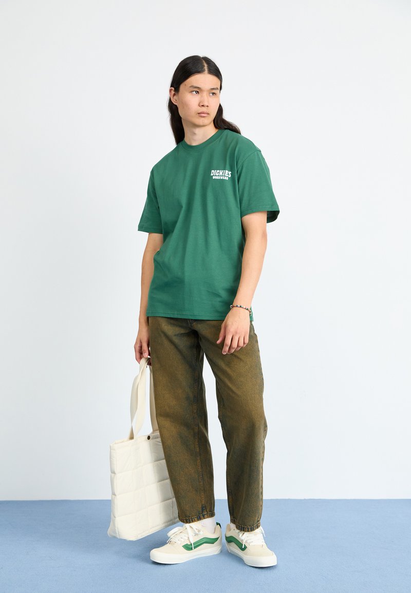 Green cotton t-shirt with logo, paired with brown baggy pants and white sneakers featuring green accents. Model holds a cream tote bag.