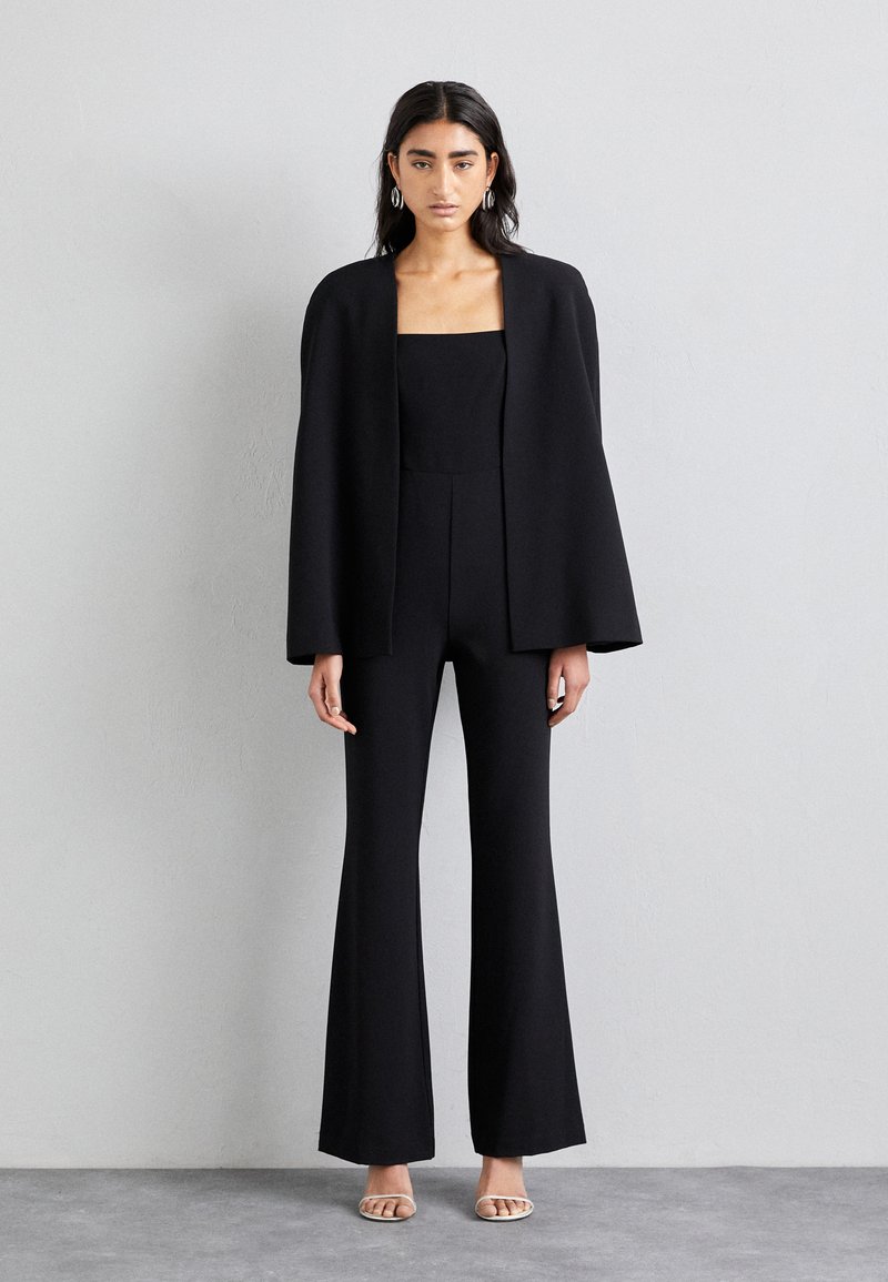 Jumpsuit nero con gambe svasate, scollatura quadrata e corpino aderente, completato da un blazer nero fluente. Tessuto liscio, hardware minimale.