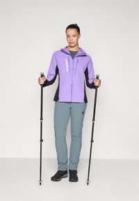 Giacca da escursionismo viola chiaro e nera con zip, pantaloni grigi, scarponi da trekking neri e bastoncini da trekking. Design elegante e funzionalità pratiche.