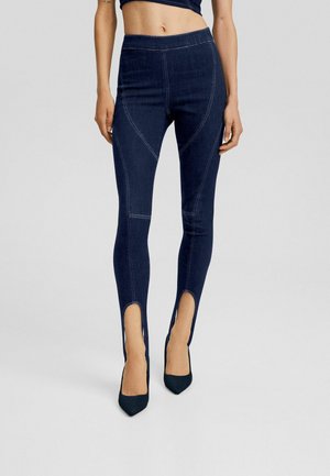 Vrouw draagt donderblauwe skinny jeans met hoge taille, voorzien van een voorkantnaad en gespleten zoom, gecombineerd met zwarte spitse hakken.