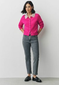 Fuchsia geripptes Cardigan mit einem weißen Spitzenkragen, Knopfleiste, getragen über grauen, hochgeschnittenen Jeans und schwarzen Loafern. Glatte Textur.