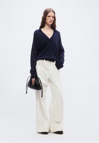 Γυναίκα που φοράει μπλε πουλόβερ V-λαιμόκοψη και λευκό παντελόνι με φαρδιά πόδια, κρατώντας μια μαύρη τσάντα με κορδόνι, στέκεται μπροστά σε απλό λευκό φόντο.