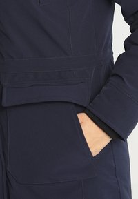 Marinblå vattentät jacka med bälte i midjan, stor ficka med klaff och en texturerad tyg för hållbarhet. Ärmarna täcker ett svart inre lager.