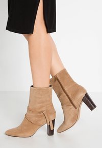 Botines de ante beige con punta afilada, detalle de correa envolvente y cremallera en la parte trasera, descansando sobre un tacón de bloque de madera oscura.