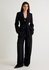 Zwarte op maat gemaakte blazer met een getailleerde taille en wijde broekspijpen. Heeft gestructureerde schouders, zijzakken en stijlvolle puntige hakken.