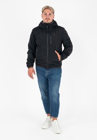 Giacca leggera nera con cappuccio, zip frontale e tasche laterali, abbinata a jeans blu e sneakers bianche. Texture liscia, design minimale.