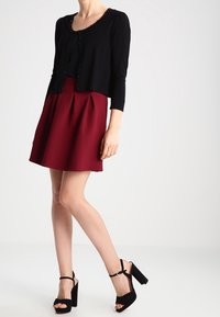 Cardigan nero corto con bordo texturizzato, abbinato a una gonna bordeaux svasata. La modella indossa sandali neri con piattaforma e cinturino alla caviglia.