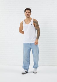 White cotton tank top with black logo, paired with light blue denim jeans. Model wearing gray and white athletic shoes, standing against a white wall.