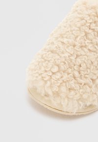 Cremefarvet fuzzy slippers lavet af tekstureret, krøllet materiale. Let afrundet form med en beige gummisål. Blød, hyggelig udseende.