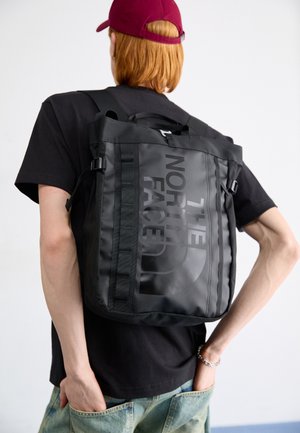 Jeune personne portant un sac à dos noir North Face avec un grand logo, un t-shirt noir, une casquette rouge et un jean bleu clair, debout devant un fond blanc.