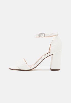 Sandale blanche à talon haut avec bride à la cheville et boucle, talon bloc et bout ouvert sur un fond clair uni.