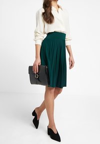 Grön plisserad kjol med hög midja, kombinerad med en krämfärgad skjorta med knappar och svarta mockaskor med spetsig tå, i handen håller en svart handväska.