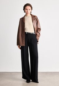 Jaqueta de couro castanha sobre um suéter bege, combinada com calças largas pretas. Um cinto preto e sapatos pretos elegantes completam o visual.