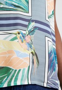 Colibrí colorido y hojas tropicales impresas en una tela estampada de color azul claro de una blusa sin mangas.
