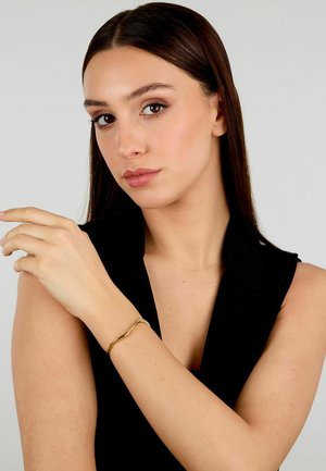 Bracciale onda in oro indossato su un polso, con un design morbido e curvo e una finitura lucida. Il modello ha la pelle chiara e i capelli scuri.
