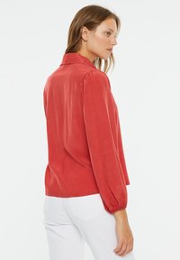 Blusa roja con cuello tipo camisa, mangas largas abullonadas y puños elásticos. Hecha de una tela suave; la parte trasera presenta un diseño sencillo y limpio.