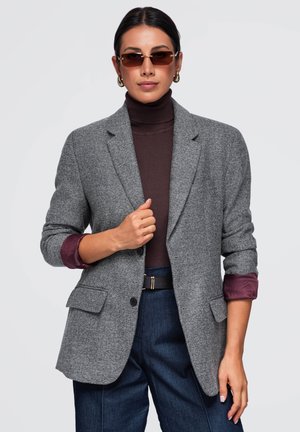 Femme aux cheveux foncés porte un col roulé marron, un blazer gris, des lunettes de soleil, des boucles d’oreilles créoles dorées et un pantalon bleu avec une ceinture noire, debout sur fond blanc.