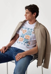Beige bomberjack over een wit T-shirt met bloemprint in oranje en teal, gecombineerd met blauwe spijkerbroek. Duidelijke plastic stoel.