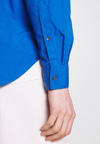 Blaues Baumwollhemd mit langen Ärmeln und geknöpften Manschetten, mit abgerundetem Saum und glatter Textur, kombiniert mit einem hellen Stoff darunter.