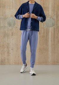Giacca di denim blu scuro con chiusura a bottoni e polsini blu chiaro a contrasto, abbinata a pantaloni della tuta viola chiaro e scarpe da ginnastica multicolori.