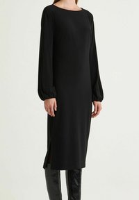 Abito midi nero con maniche lunghe e leggermente gonfie e scollatura rotonda. Presenta spacchi laterali e tessuto liscio, abbinato a stivali neri lucidi al ginocchio.