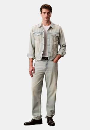 Jonge man draagt een licht gewassen spijkerjack en spijkerbroek met een wit T-shirt en zwarte nette schoenen, staand met één hand in de zak.