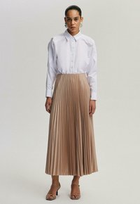 Valkoinen paita nappisuljenta, ja pitkä beige pleatattu hame. Mallilla on hopeiset korvakorut ja hihnalliset beige korkokengät. Sileä kankaan pinta.
