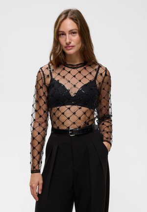 Femme aux longs cheveux bruns porte un haut noir transparent avec un motif de diamants perlés sur un bralette noir et un pantalon noir taille haute avec une ceinture.